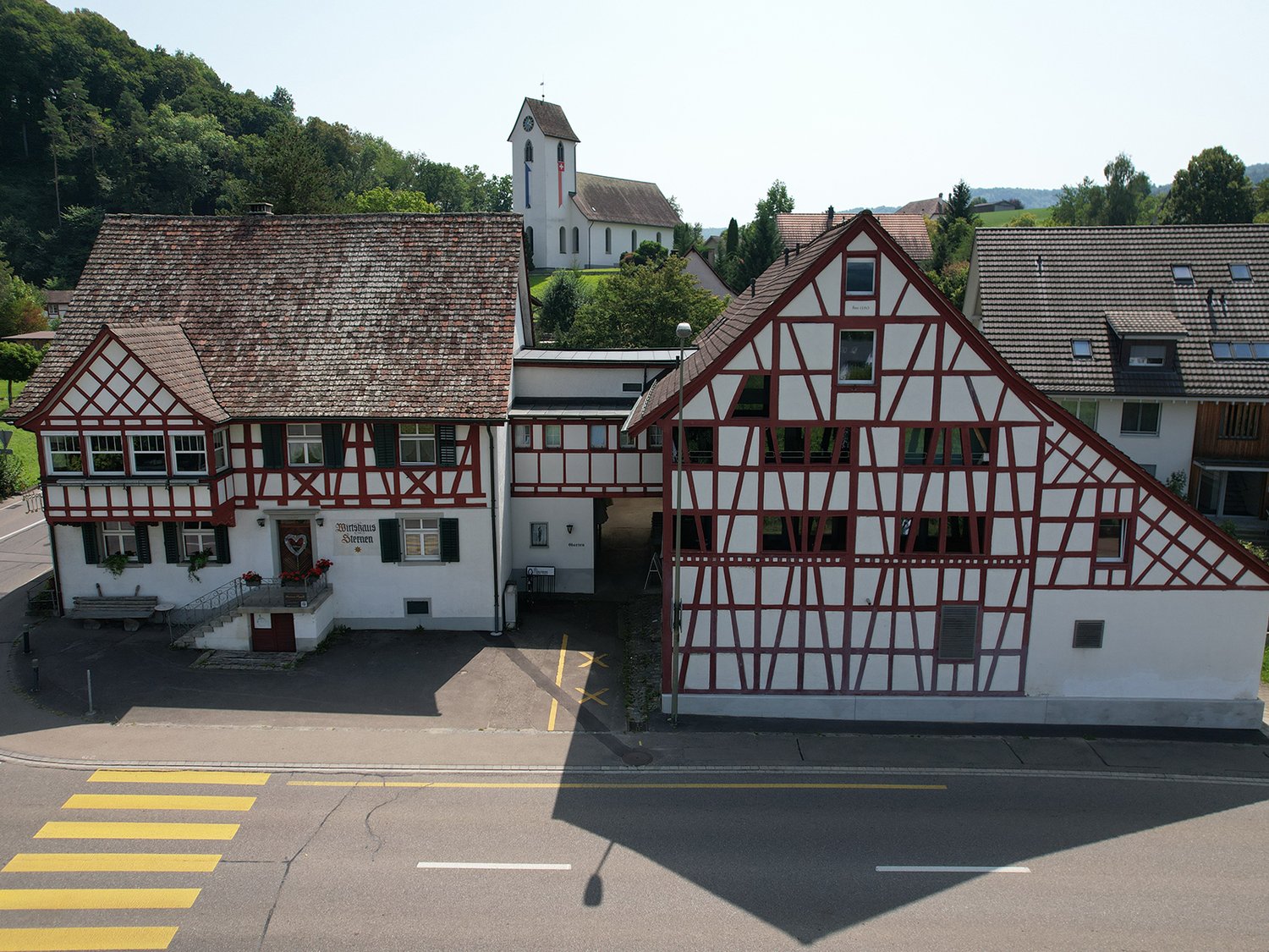 Building with a traditional design, two stories, brown and white checkered exterior, large windows, entrance with a staircase, sign reading 'Wurschaus Sormen', small wooden bench, church with a steeple in the background, green surroundings