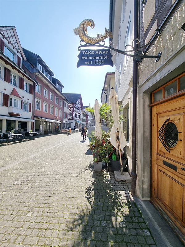 a cobblestone street, people walking, TAKE AWAY sign, outdoor dining tables, potted plants