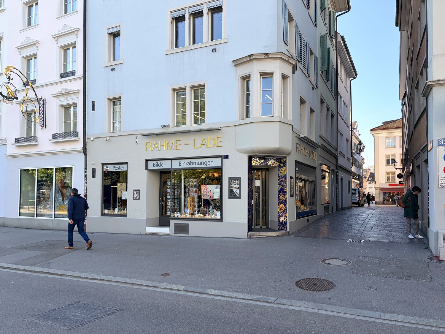 A three-story building with a shop on the ground floor, featuring a sign that reads 'RAHME-LADE'. The shop has large glass windows displaying various items. The building has a modern architectural design with white walls and green window shutters. There a