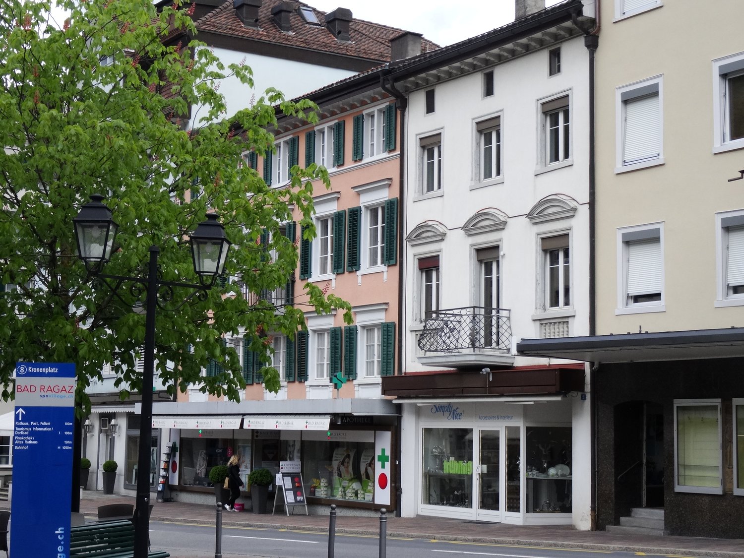 Three buildings with different colors, one with a balcony, shops on the ground floor, street lamp, tree, and bench