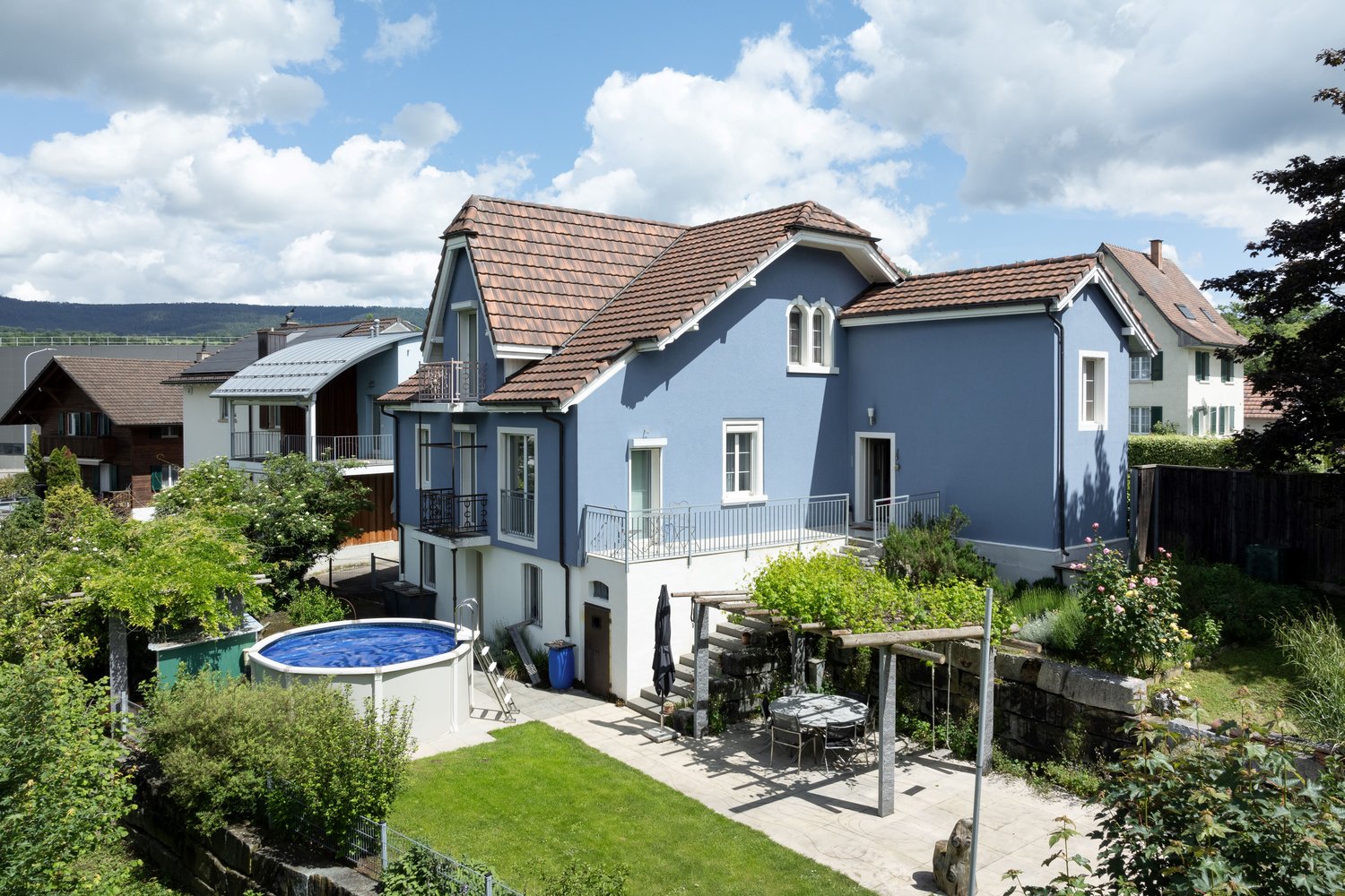 Blue house with brown roof, swimming pool, garden, terrace with table and chairs, wooden pergola, plants, fence