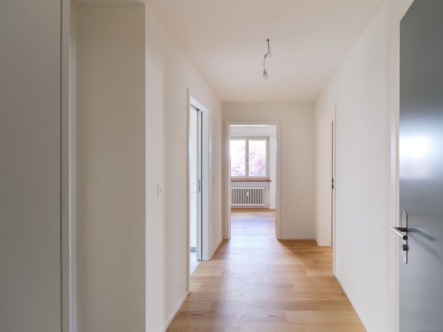 light colored wooden floors, white walls, radiator, door, window, light fixture