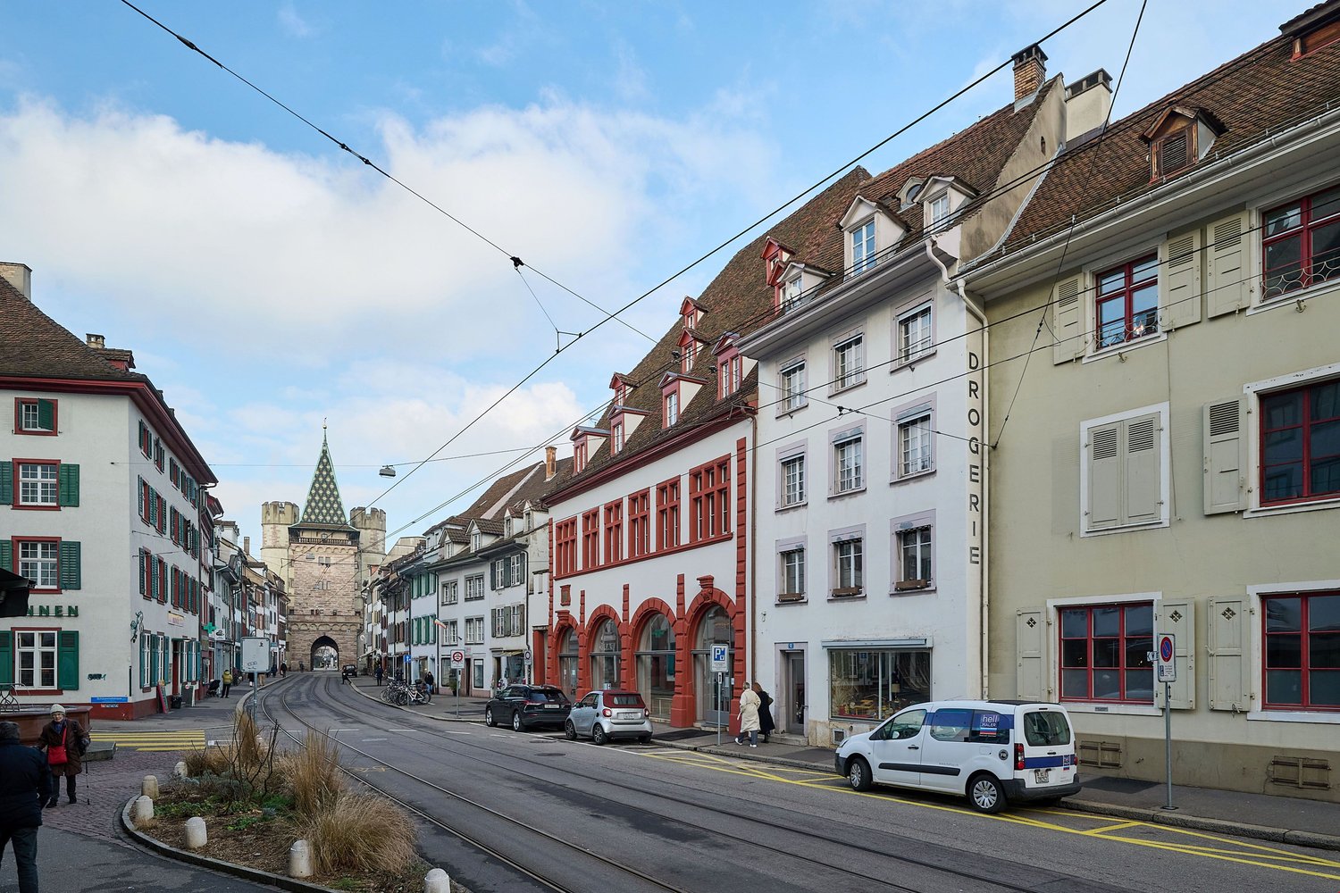 Street with buildings, cars parked on the side, pedestrian sidewalk, people walking, street lights, tram tracks
