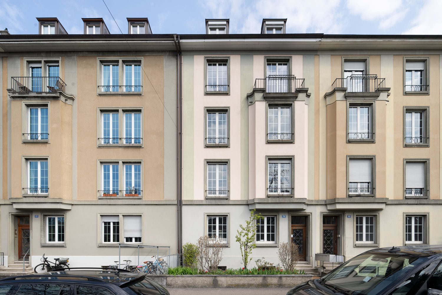 5-story building, beige and white exterior, multiple windows, balconies, front garden with plants and bicycles