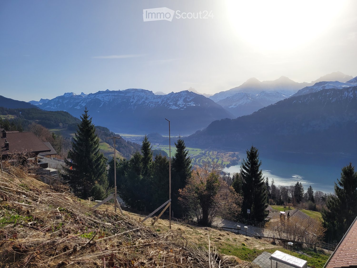 Aerial view of mountainous terrain with lush vegetation, a serene body of water, distant houses, and a clear sky.