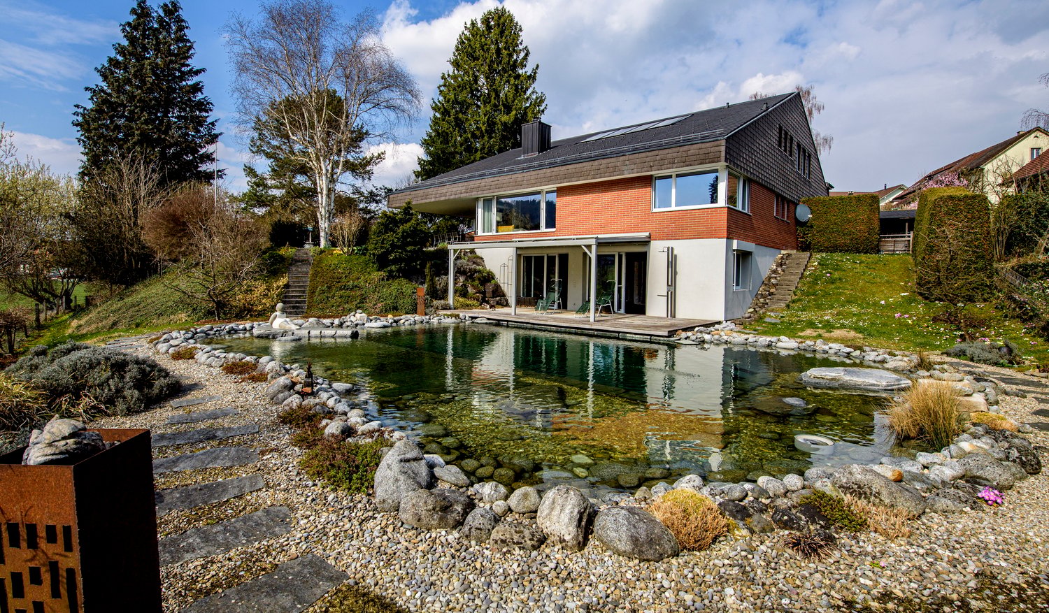 Modern house with swimming pool, brick and white walls, solar panels on the roof, surrounded by a garden with rocks and plants