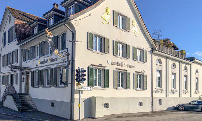 A multi-story building with a traditional European architectural style, featuring a light-colored facade, green shutters, and a prominent sign on the front. The building appears to be a commercial or residential property located in an urban setting.