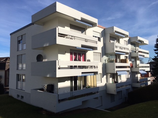 A 3 story white apartment building with multiple balconies, windows, and a roof terrace.