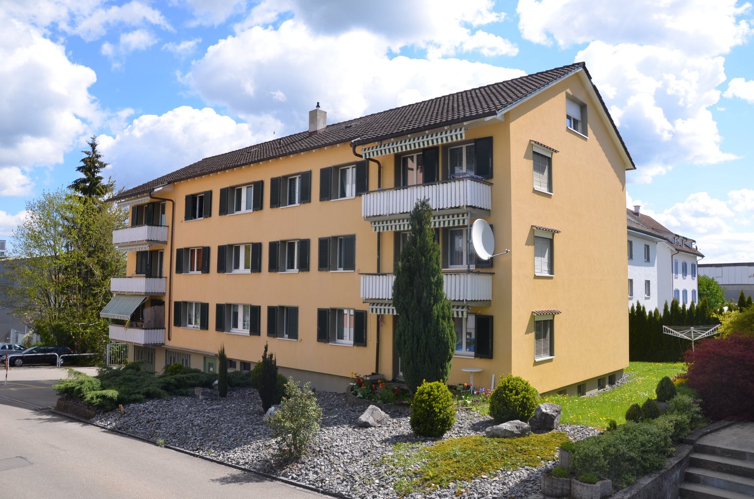 3-story apartment building, yellow paint, brown roof, white railings on balconies, multiple windows, satellite dish on the side