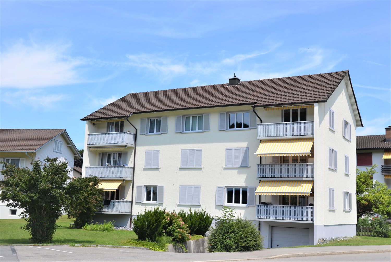 3 story apartment building, white walls, brown roof, yellow window shutters, balconies, 2 garages