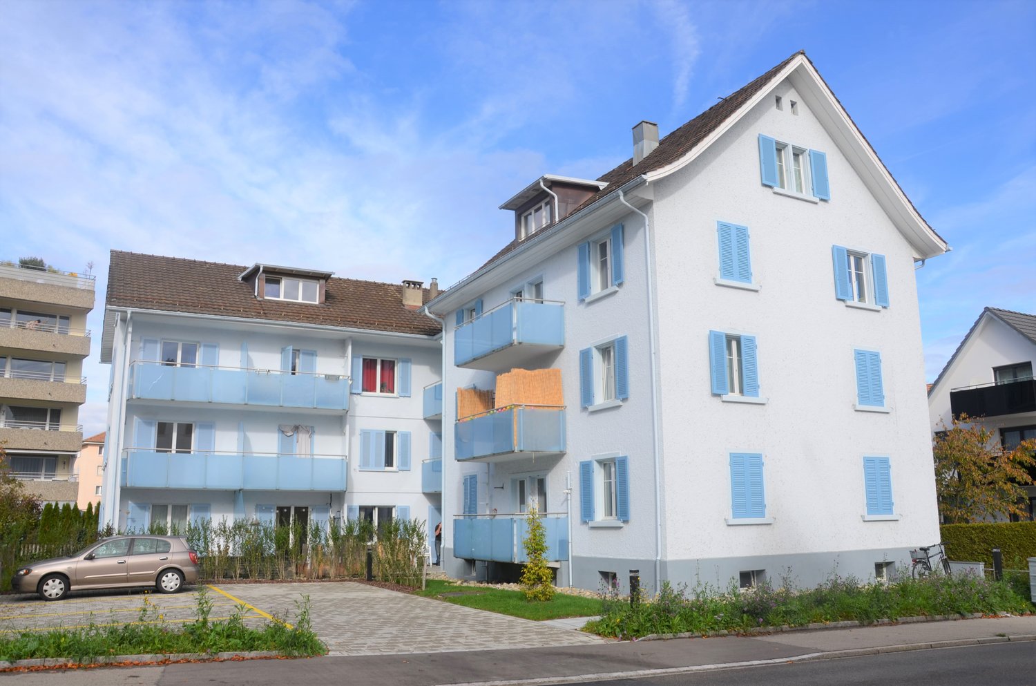 Multi-story apartment building with white exterior, blue shutters, and balconies. There is a parking area in front of the building.