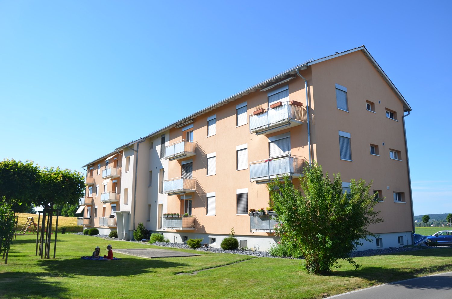 5 story apartment building, beige color, multiple balconies, garden in front, parking lot, car in the picture
