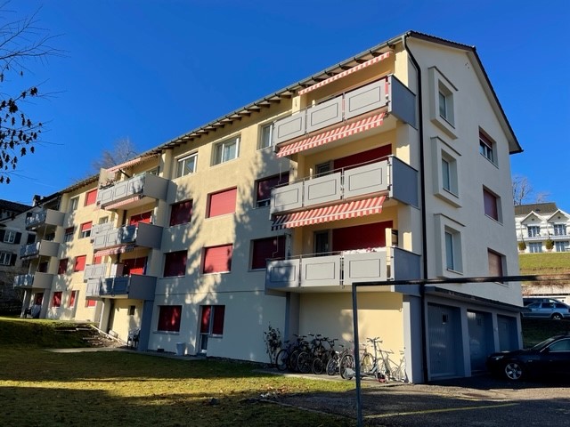 Modern 4-story apartment building with balconies and garages. Balconies have striped awnings. Bicycles are parked in front of the building. A car is parked in the garage. There is grass and plants in the foreground.