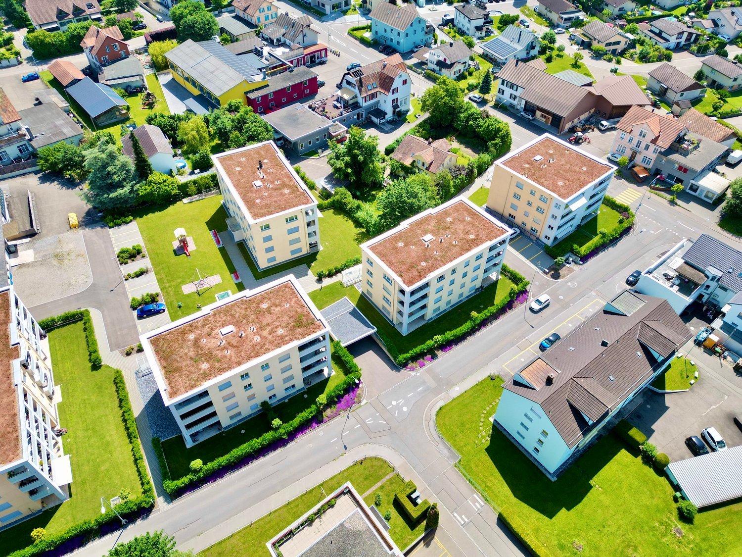 Group of apartment buildings, surrounding houses, green lawns, roads, trees, parking areas, cars parked.