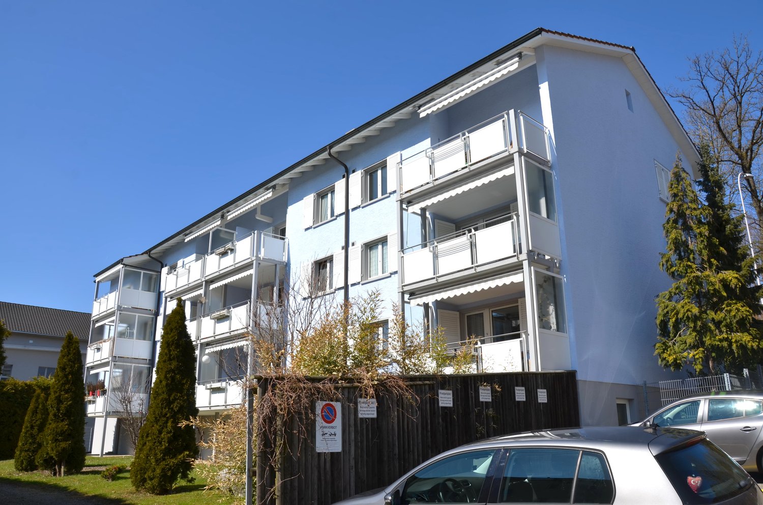 Multi-story apartment building, white exterior, multiple balconies, several cars parked in front, greenery around the building