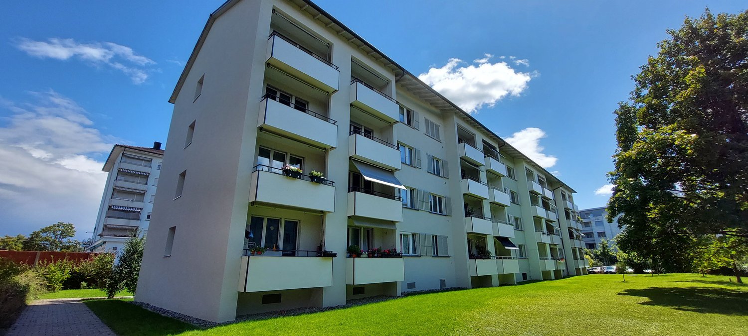 Large multi-story apartment building with balconies, large grassy lawn, several trees, other buildings visible in the background