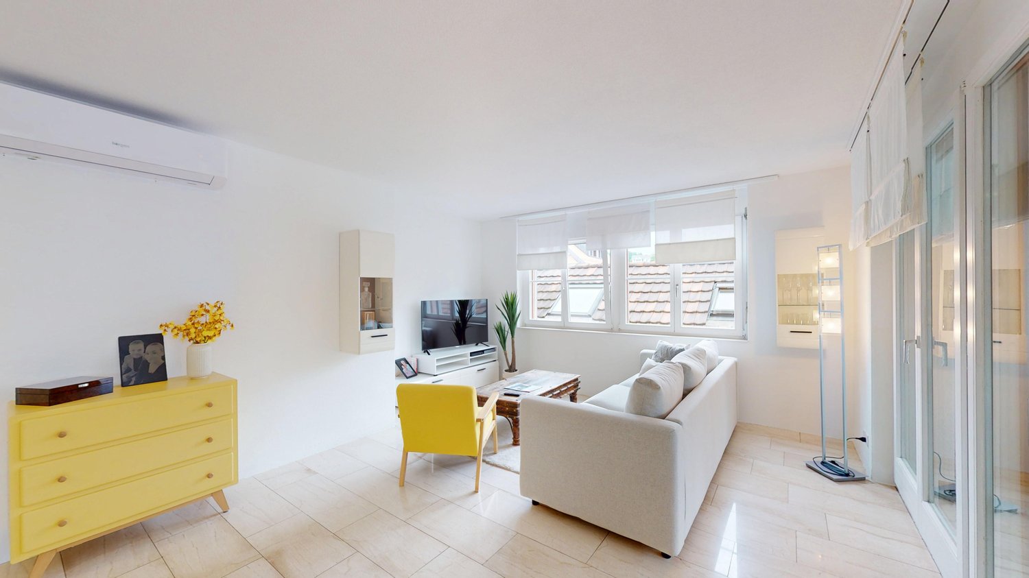 Bright and airy living room with large windows, white walls, hardwood floors, a white sofa, a yellow accent chair, a TV stand, and a yellow dresser with decor items.