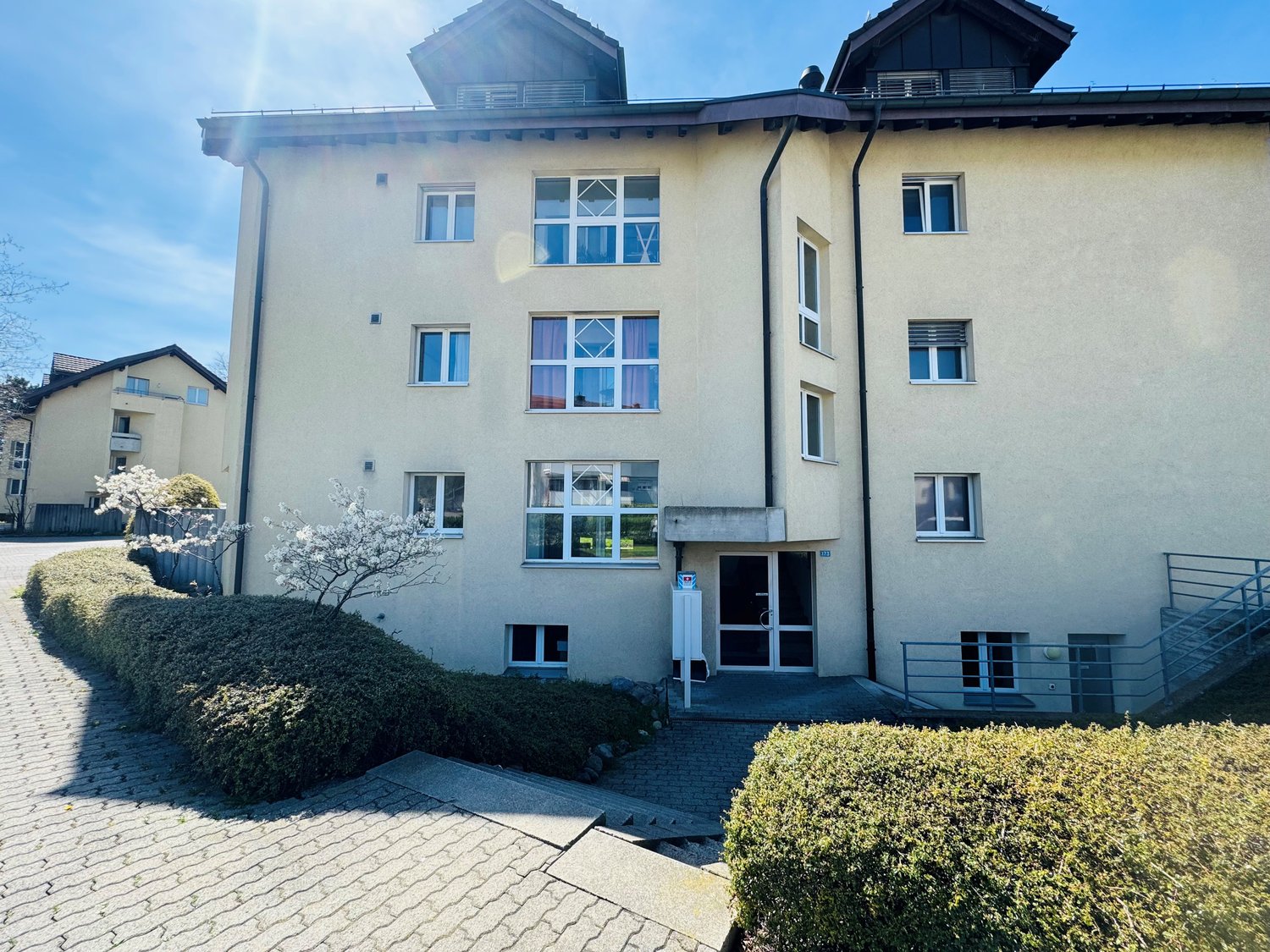 3 story apartment building, yellow facade, multiple windows, entrance with double doors, small trees and bushes, brick path