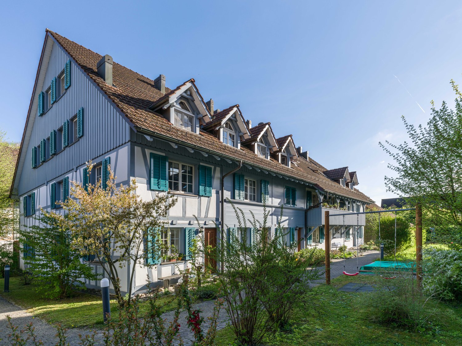 Traditional Swiss house, multiple levels, brown roof, blue shutters, large windows, raised ground floor, garden, swing, play equipment