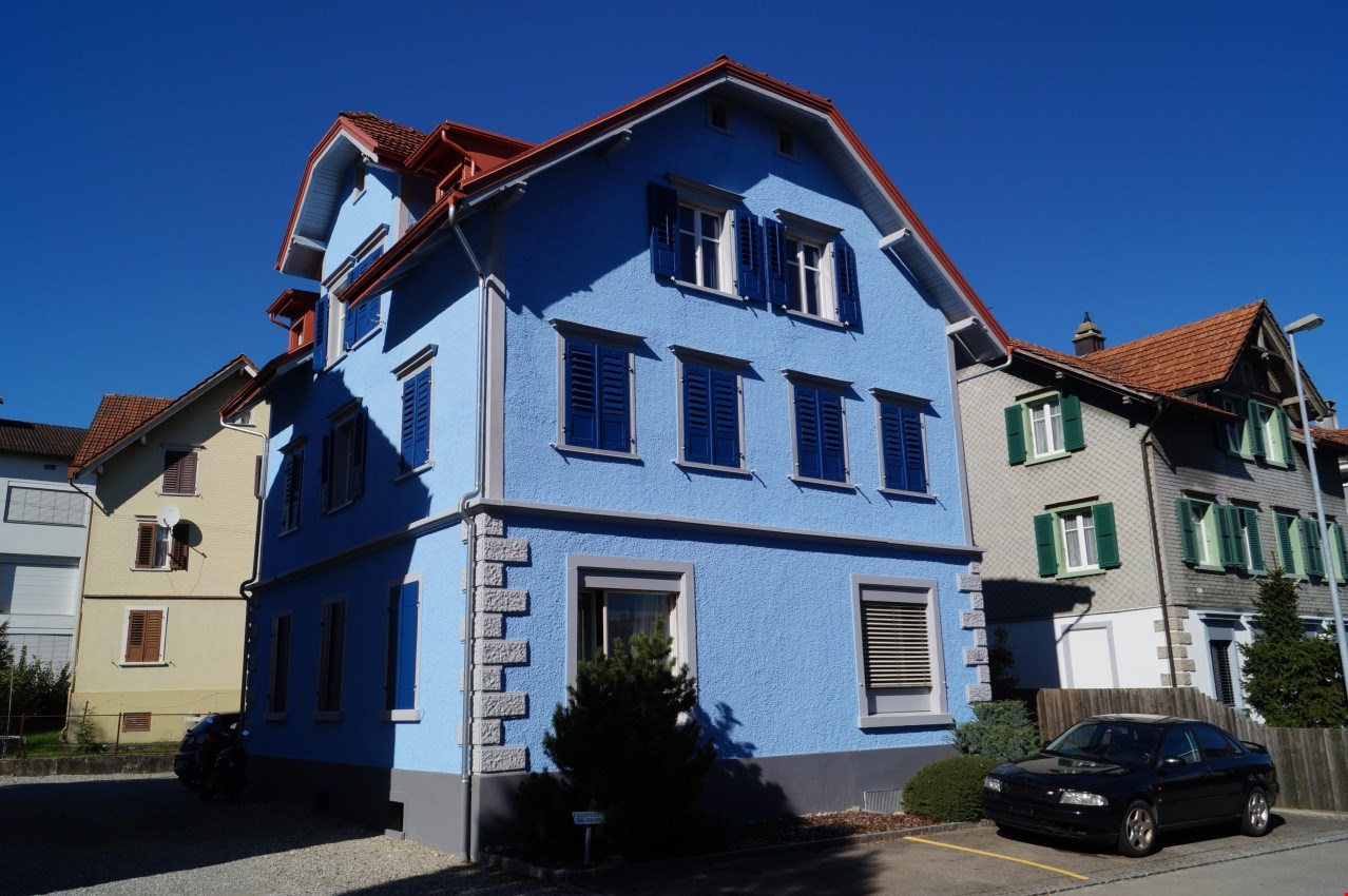 raised ground floor, blue exterior walls, red roof, two cars parked in front, green window shutters