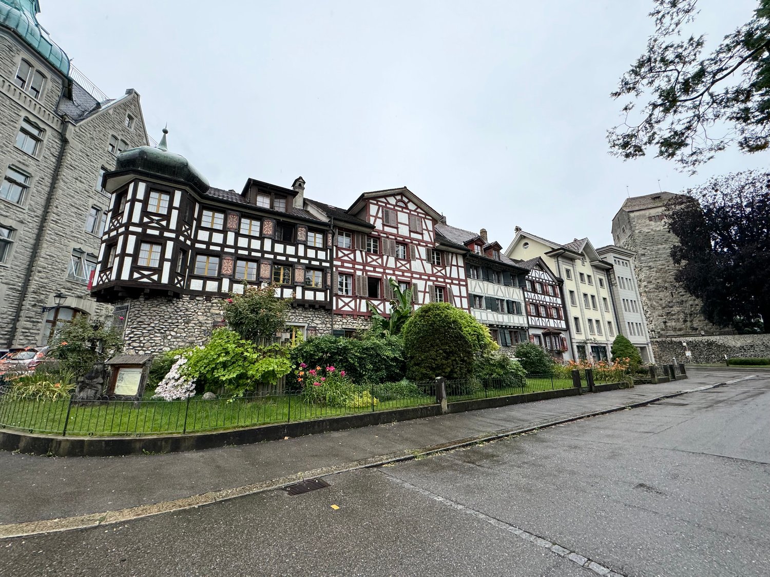 Traditional architectural style building, 3 stories, stone and timber facade, dormer windows, steep roofs, greenery, paved street