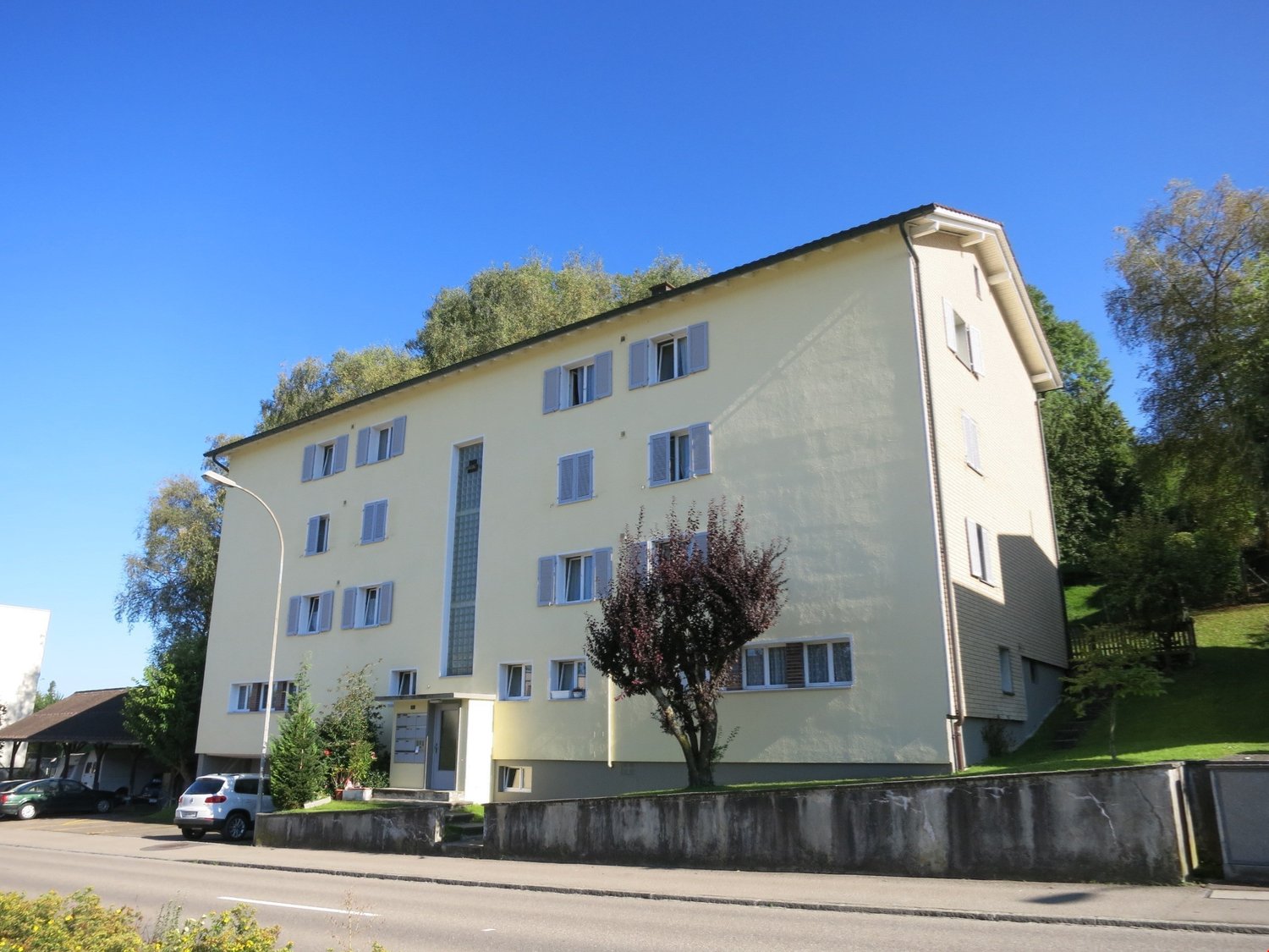 3 story yellow building, white shutters, entrance gate, concrete fence, car parked, trees