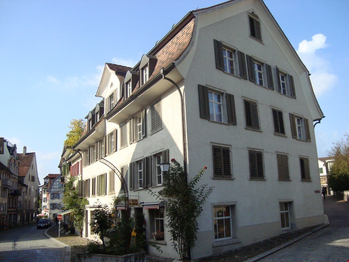 3-story building with a slanted roof, white exterior walls, and black shutters on the windows. The building has a traditional European architectural style and is located on a street with other similar buildings.