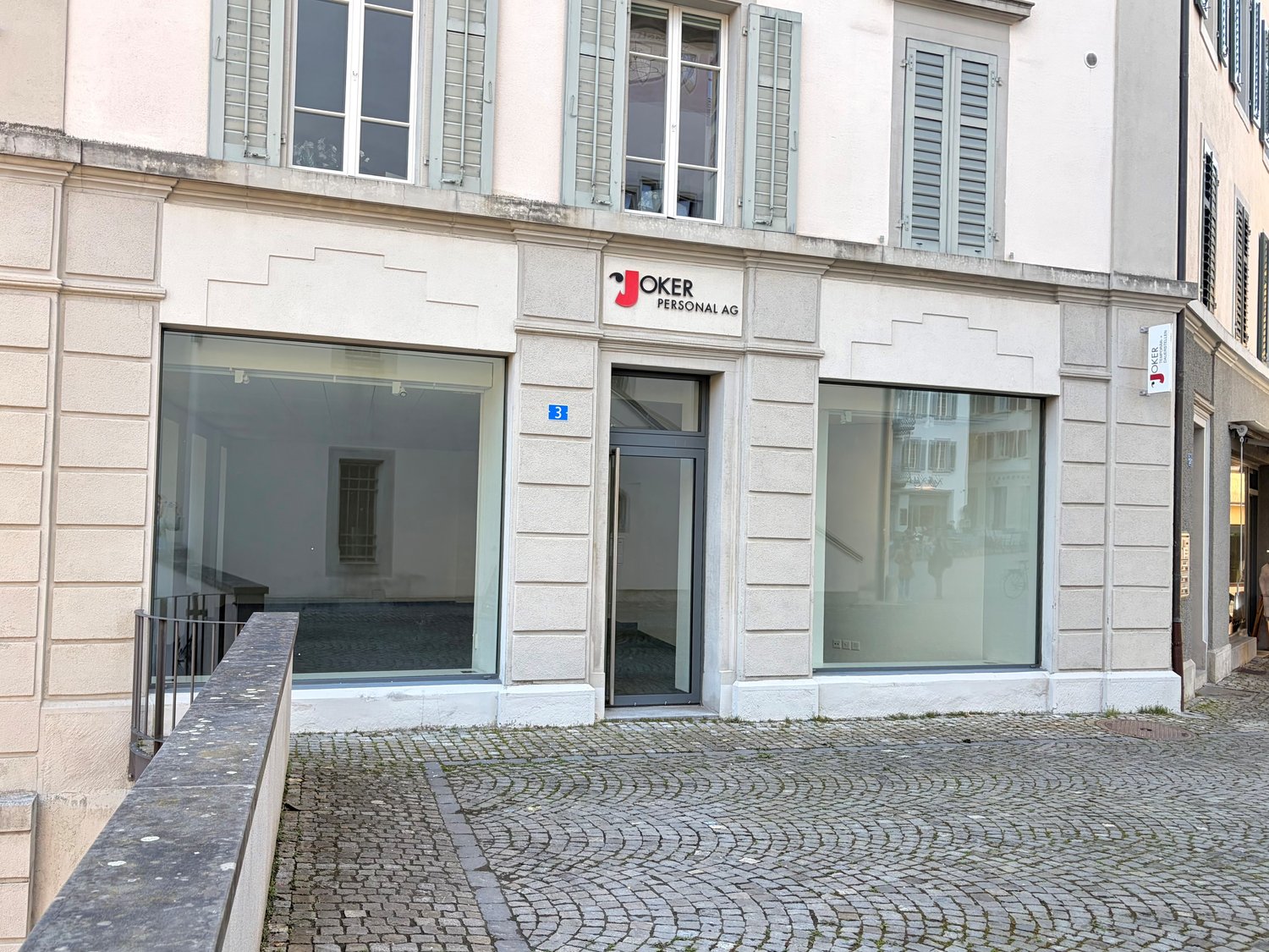 Facade of a building with glass windows and doors, a sign reading 'Joker Personal AG', and a cobblestone pavement in front.