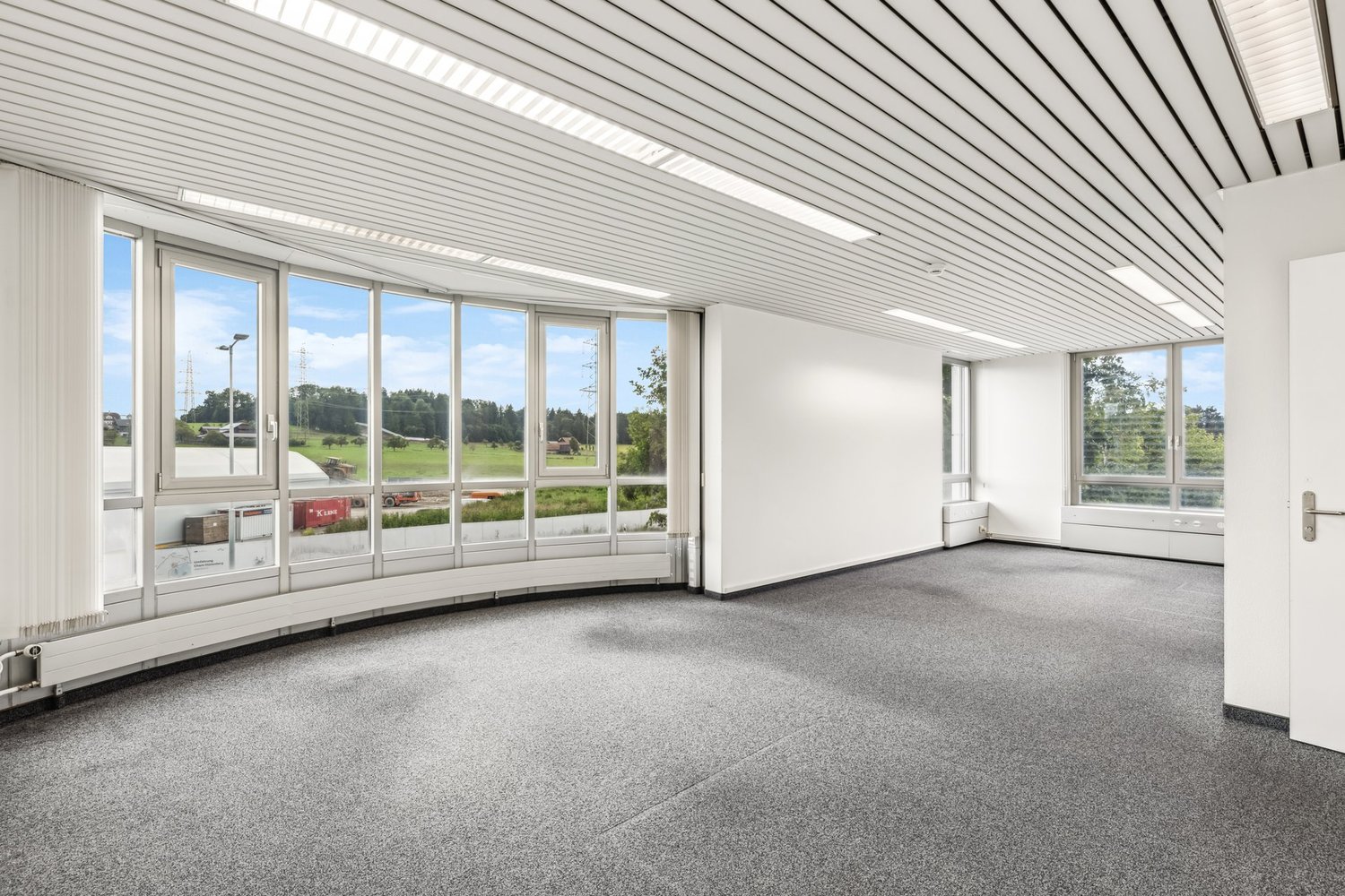Large open office space with floor-to-ceiling windows providing a view of the surrounding landscape. The space has a white ceiling with recessed lighting and a gray carpeted floor. The walls are white and there is a small storage area visible in the corne
