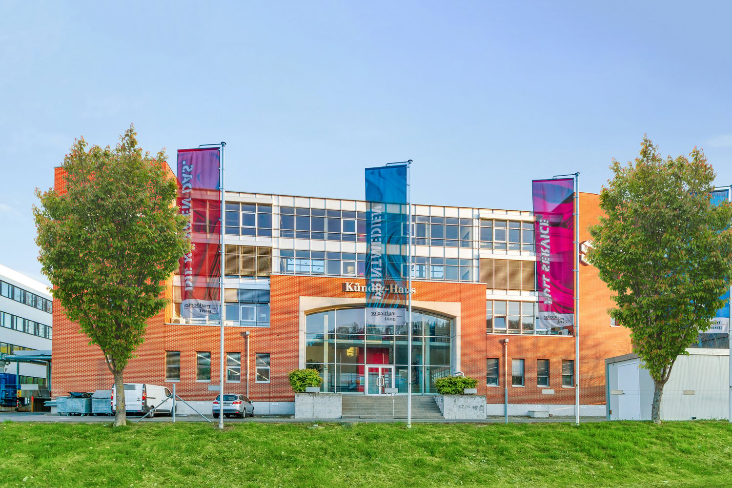 modern office building, red brick, glass windows, three flags, small front yard with grass, cars parked in the side