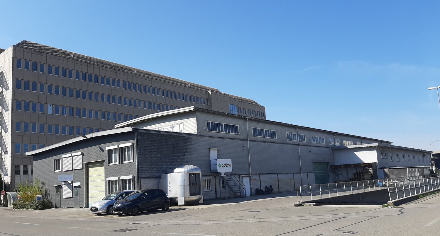 Two-story building with grey exterior, two cars parked, multiple windows, two doors, staircase at entrance, sign reading 'Optumo'.