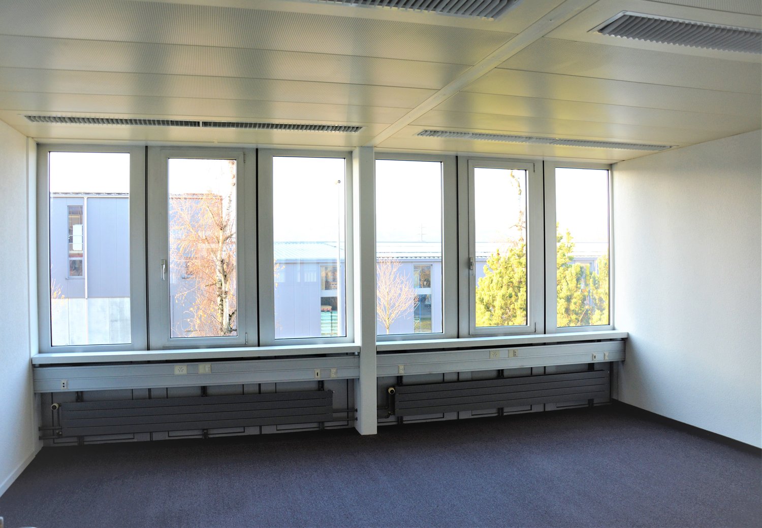 An empty room with large windows, radiators, and carpeted floor.