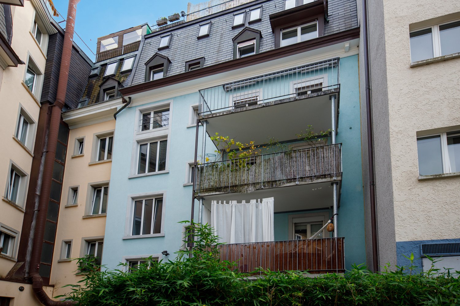 multi-story building, blue and white walls, multiple windows, two balconies with wooden railings and plants