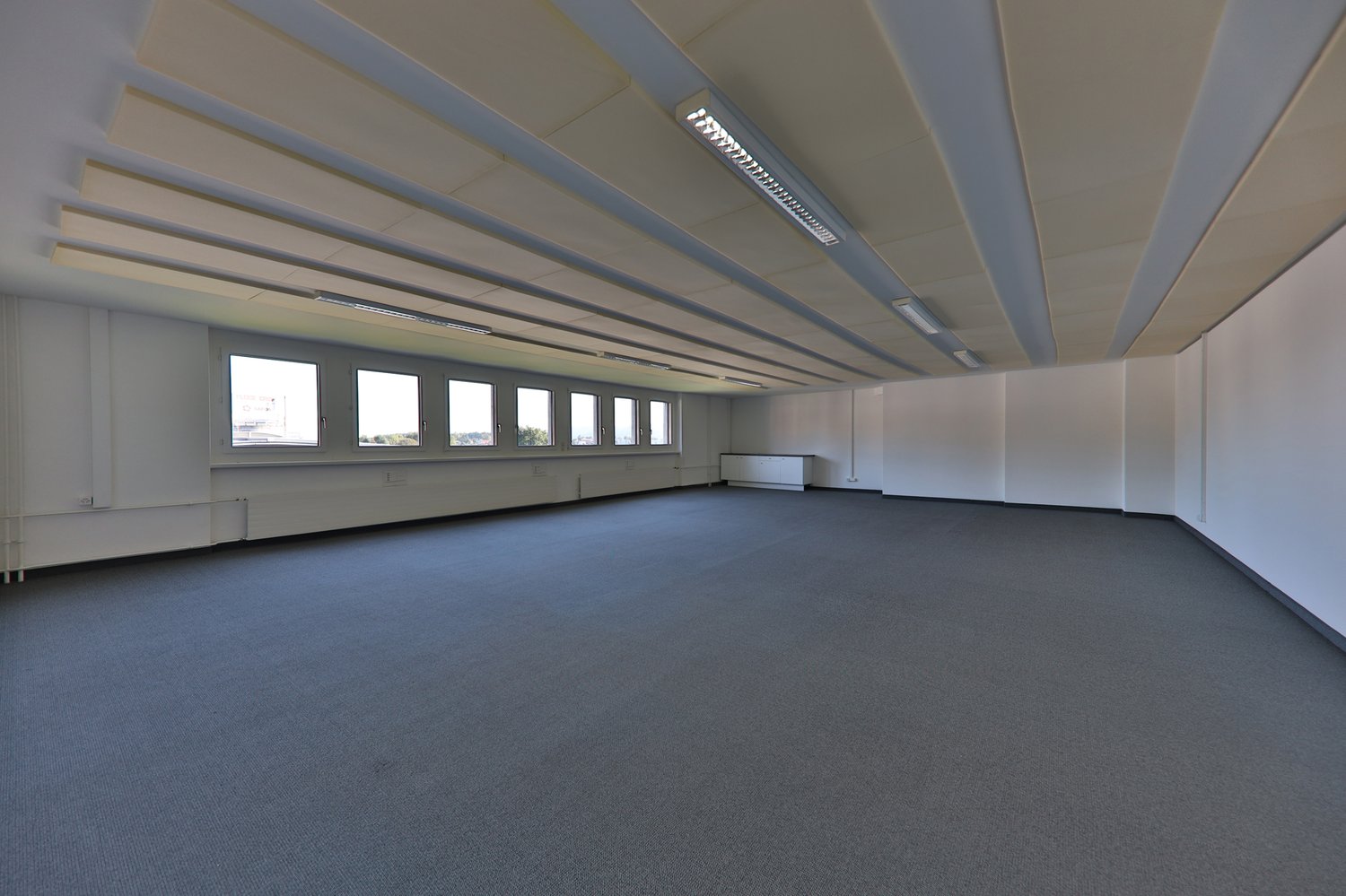 Large open office space with high ceilings, multiple windows, and a carpeted floor. The room appears to be empty and ready for use.