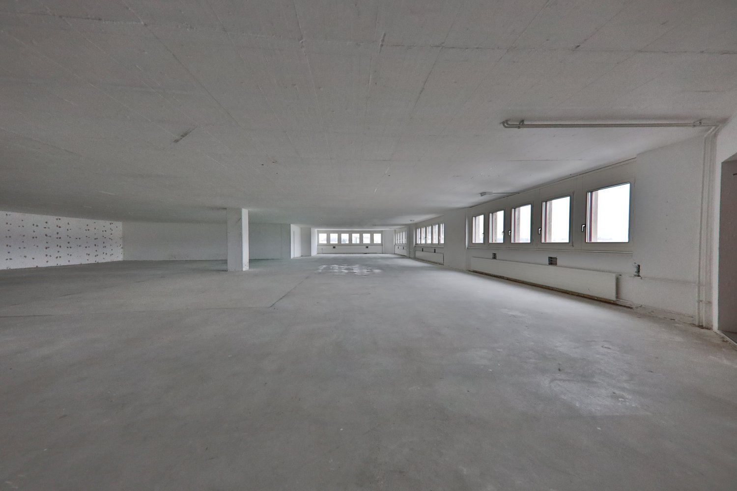Large open space with concrete floor, white walls, and multiple windows along the sides. The space appears to be an unfinished commercial or industrial building, possibly a warehouse or factory.