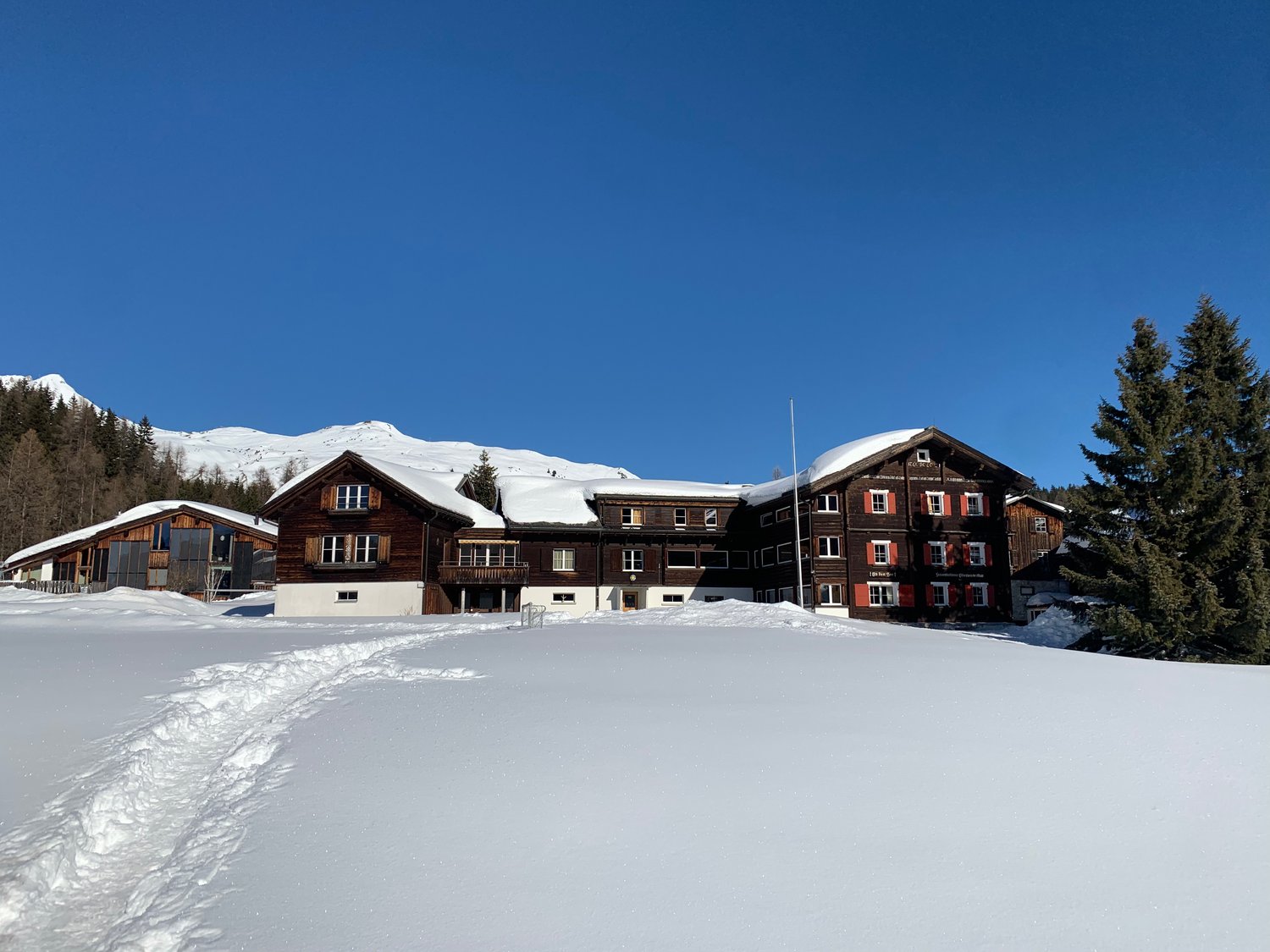 A wooden building covered in snow with several windows, two smaller houses on the left, trees in the front, mountains in the background