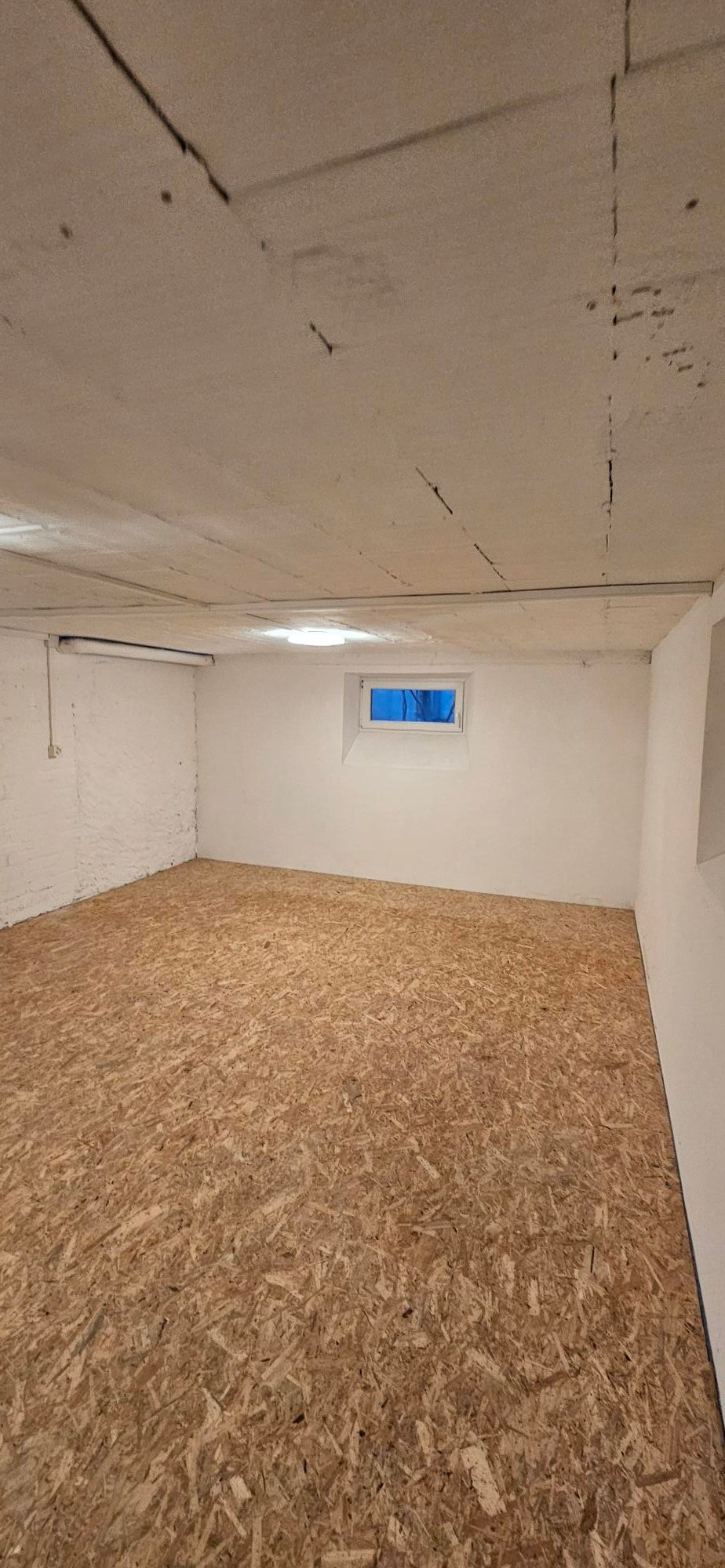 unfinished room with white walls, unfinished ceiling with cracks, small window, unfinished floor with plywood