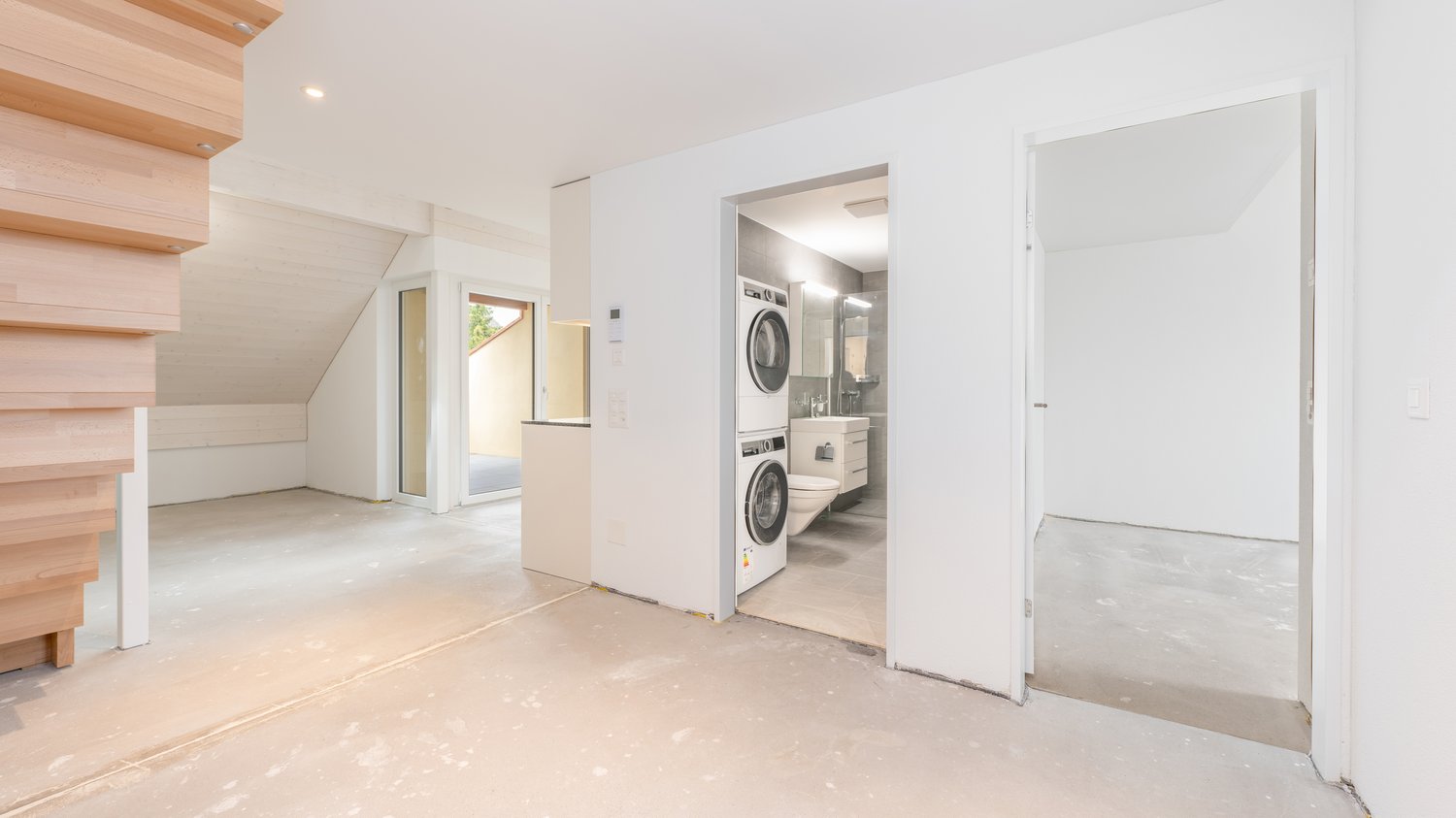 Unfinished attic space with exposed wooden beams, white walls, and a laundry room visible through the open doorway containing a washer and dryer.
