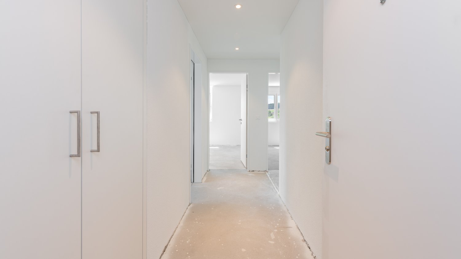 Long, narrow hallway with white walls, recessed lighting, and a white door at the end. The floor appears to be a light-colored tile or laminate material.