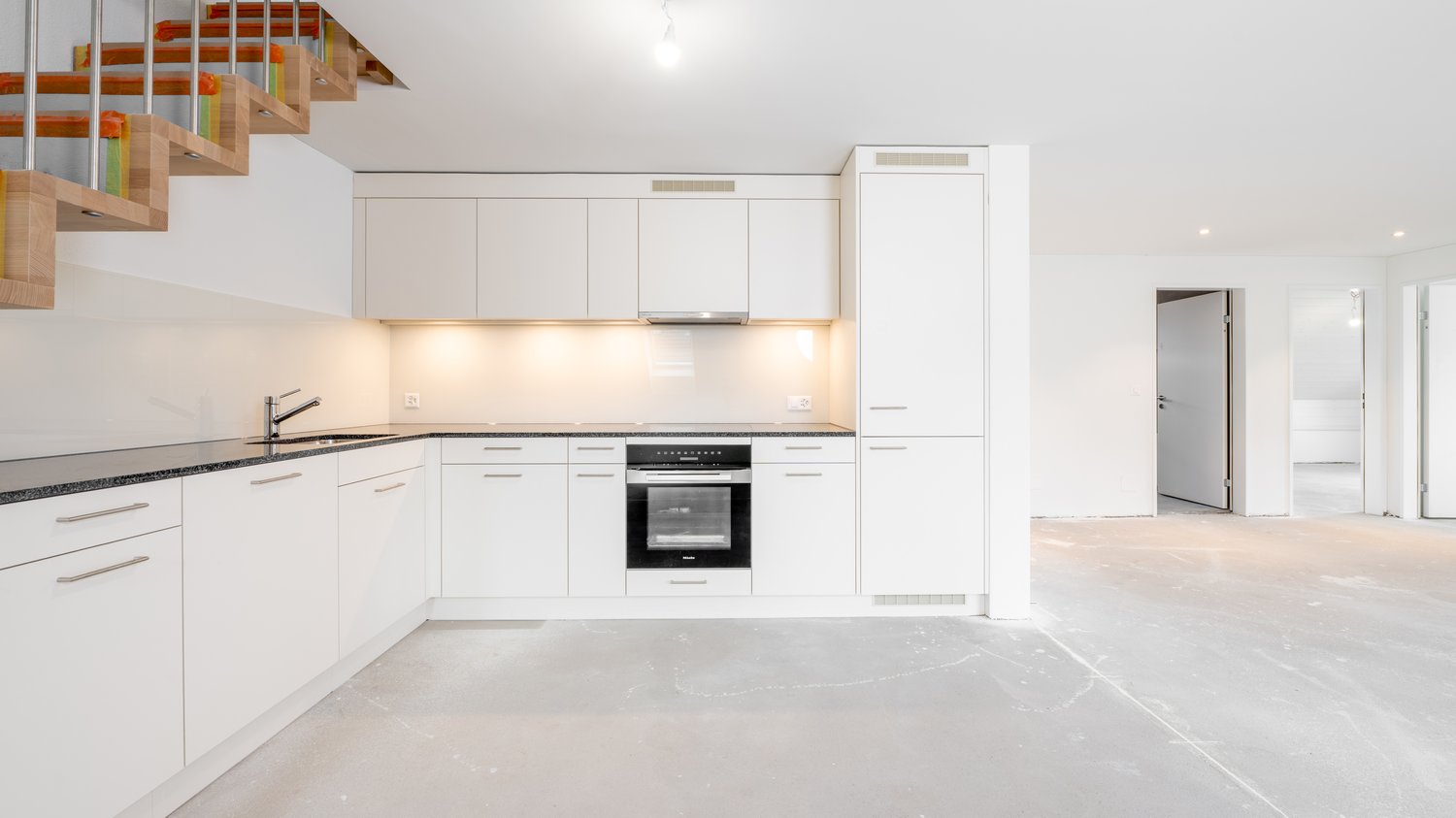 Modern, open-concept kitchen with white cabinets, black countertops, built-in oven and dishwasher, and hardwood stairs leading to an upper level