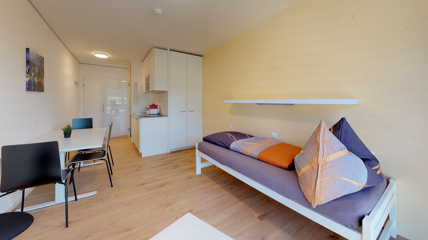 The image shows a studio apartment with a kitchenette, a dining area, and a sleeping area. The apartment has hardwood floors, white walls, and large windows. The sleeping area features a bed with colorful bedding and pillows. There is a wardrobe and a she