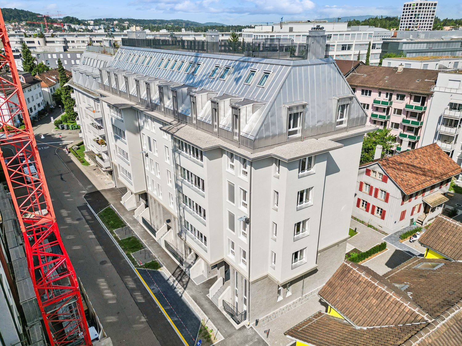 A modern white apartment building with a grey roof, multiple windows, and a metallic exterior. It is situated among other buildings in a city setting.