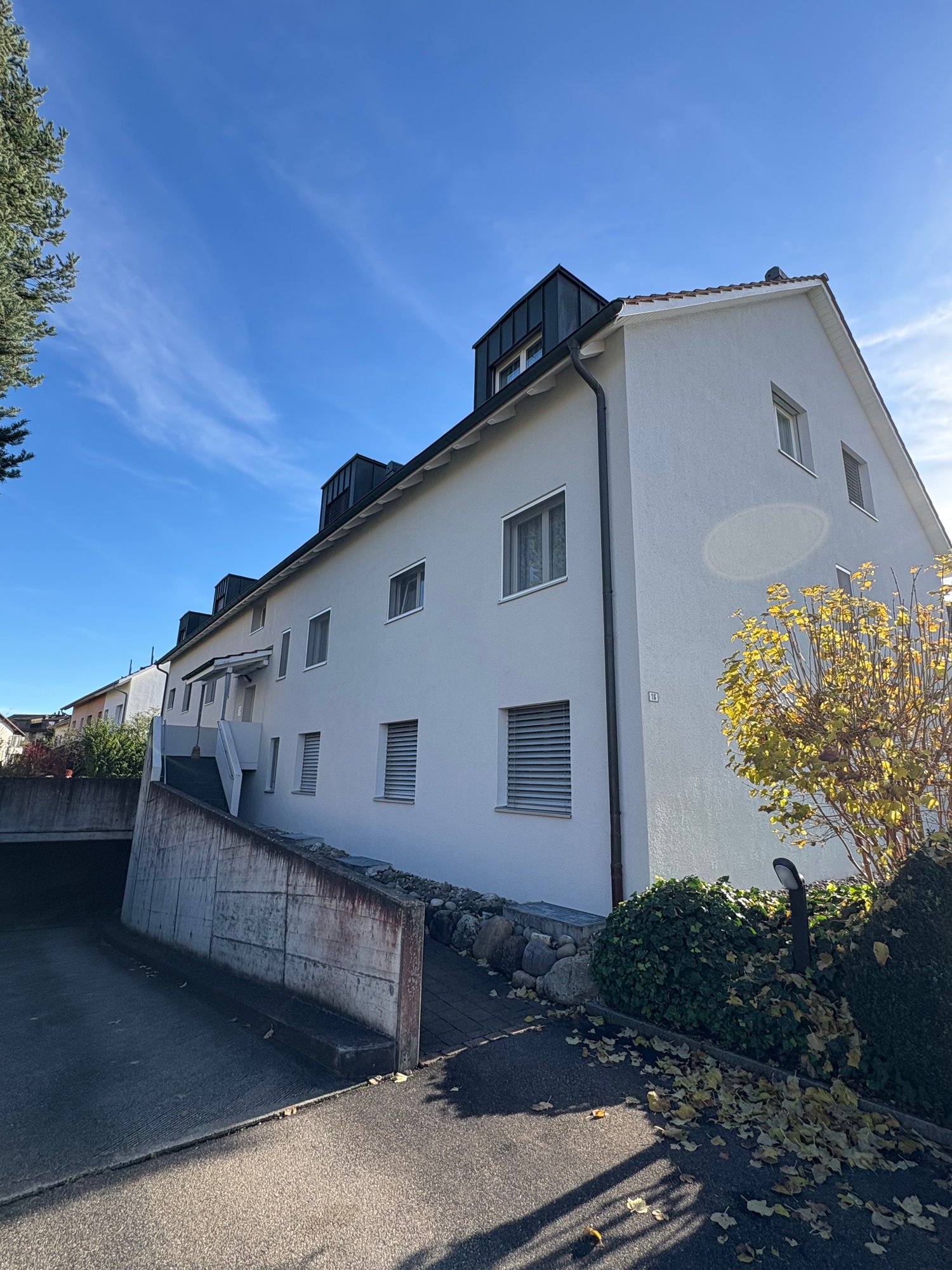 White apartment building with multiple windows, a concrete wall with stairs, an underground parking area, and trees in front.