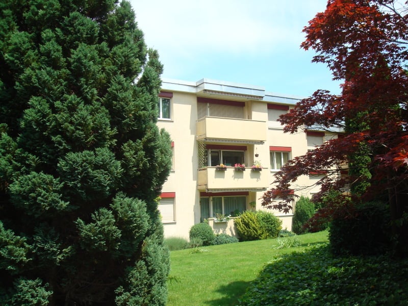A three-story yellow building with balconies and windows. There are trees and shrubs in front of the building. The balconies have potted plants and flowers. The building is surrounded by a green grassy area.
