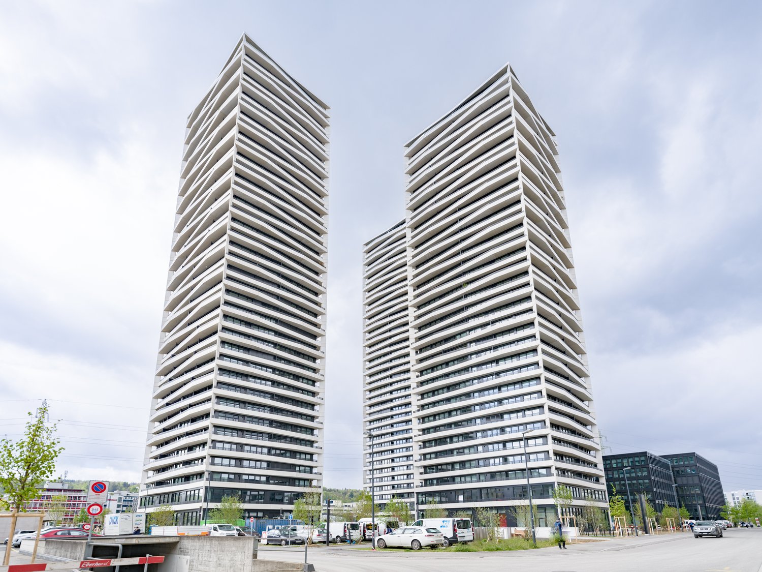 Skyscraper building, two tall buildings, concrete exterior, many windows, car park in front, street, trees, other buildings