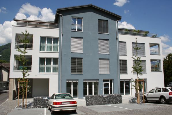 White and blue apartment building, 4 stories, garage, car parking spots, trees