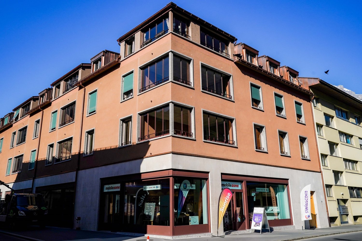 Apartment building with modern architecture, multiple floors with many windows, storefronts on the ground floor, balcony on top floor, located in an urban setting