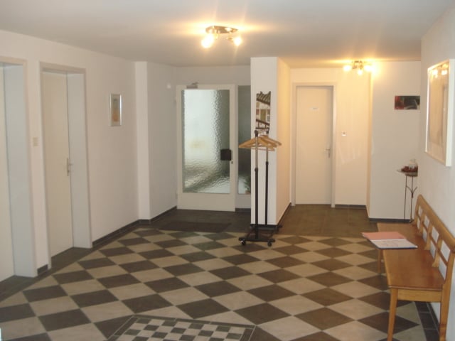 White walls, checkered flooring, wooden bench, glass door, white doors, hanging coat rack, picture frames on wall