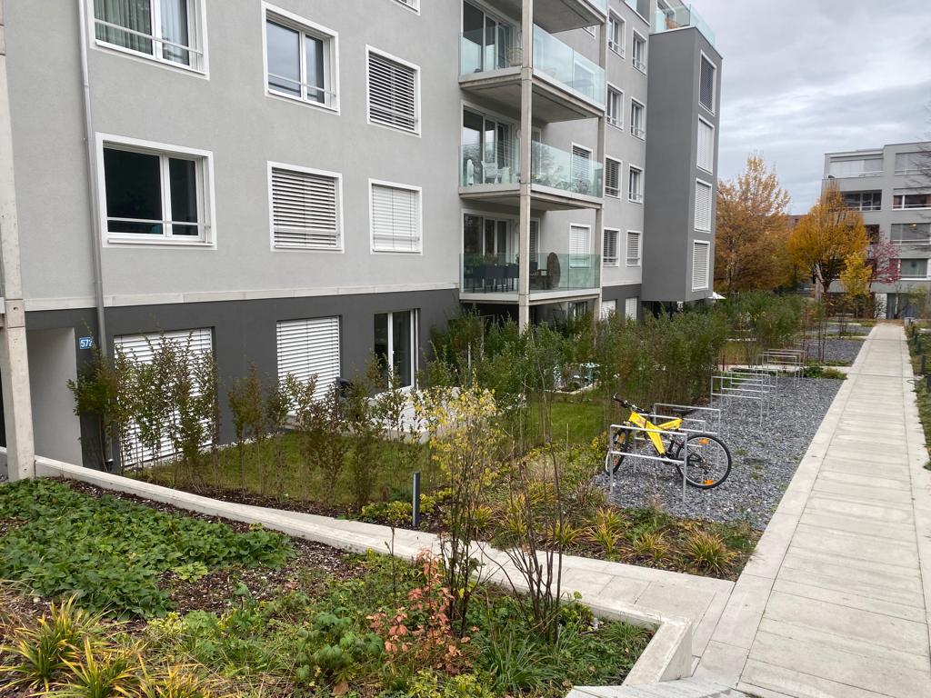 Gray exterior, multiple windows, balconies, entrance walkway, bike parking, landscaped area, and a neighboring building