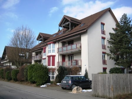 An apartment building with multiple floors, brown roof, red trims, several balconies, parking area, trees, and plants